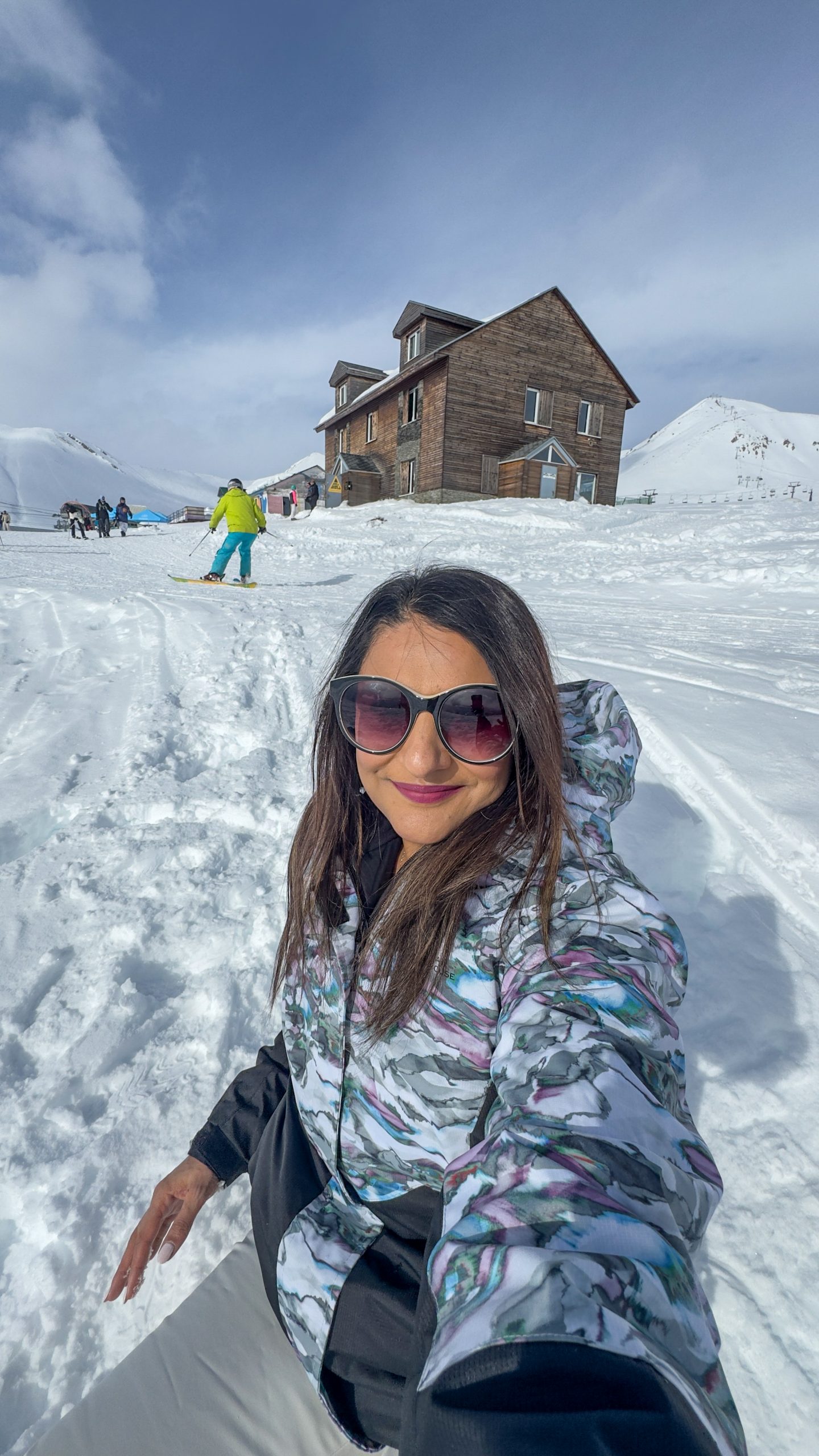 Woman sitting in the snow in Gudauri wearing sunglasses and smiling at the camera with a building and skiers behind her