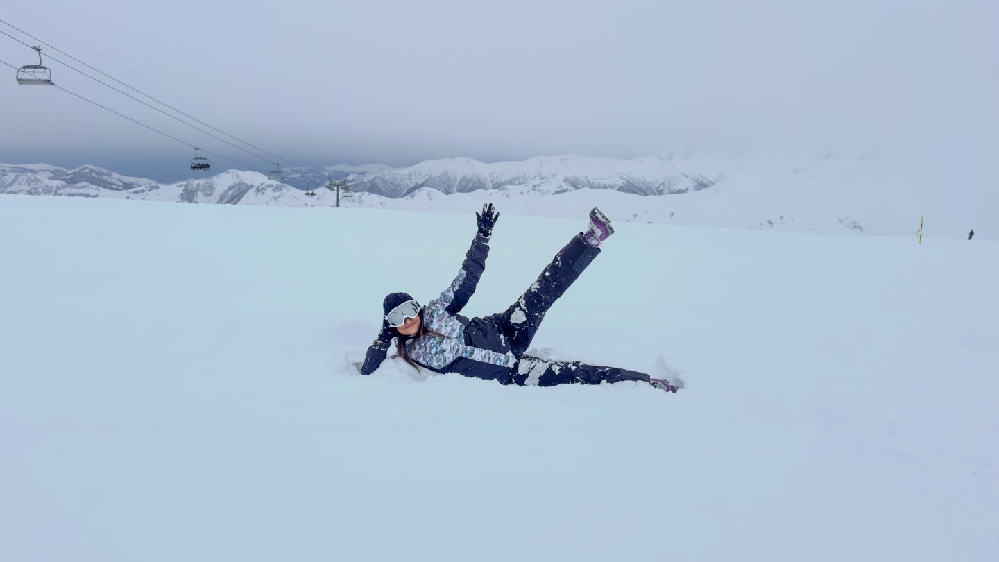 Woman in ski gear lying in the snow in the mountains with one leg up ready for her first time skiing in Gudauri