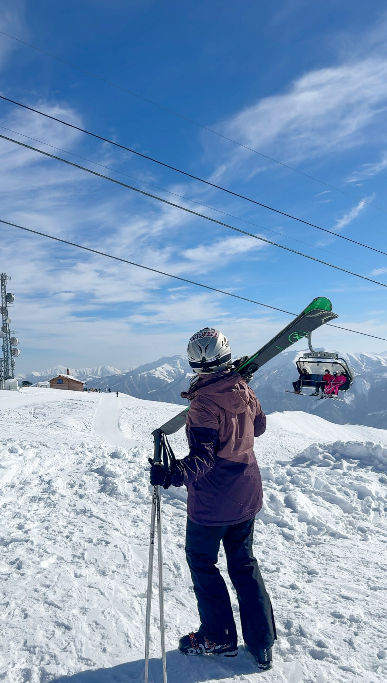 Girl carrying ski gear over her shoulder in the snow with a chair lift in the background