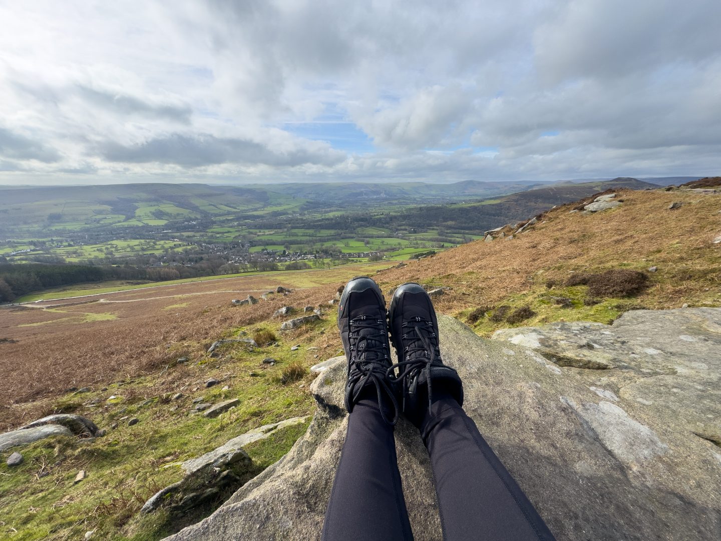 A woman's legs showing her wearing hiking boots sitting on the edge of a cliff looking at the rolling hills of Hope Valley