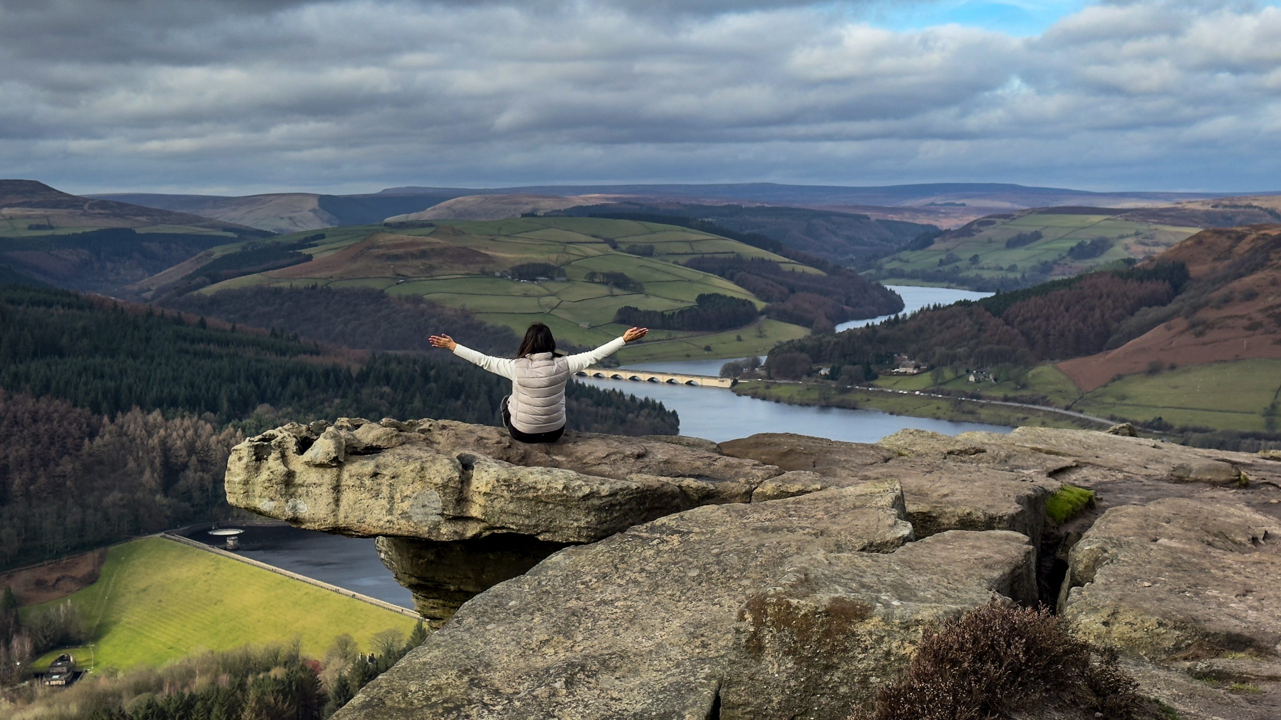 A woman sitting on the edge of a cliff at Bamford Edge overlooking the Ladybower Reservoir during a Peak District walking and wellness break.