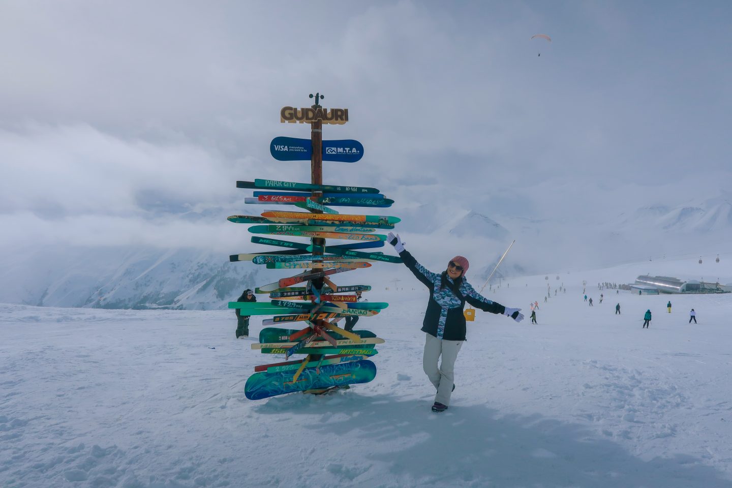 A woman standing in the snow in the mountains with a sign showing she is in Gudauri