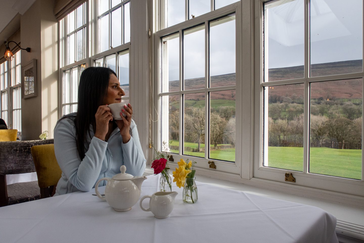 A woman sitting at a Breakfast table at Losehill Hotel and Spa with a cup of tea and looking out to the rolling hills during a Peak District walking and wellness break.