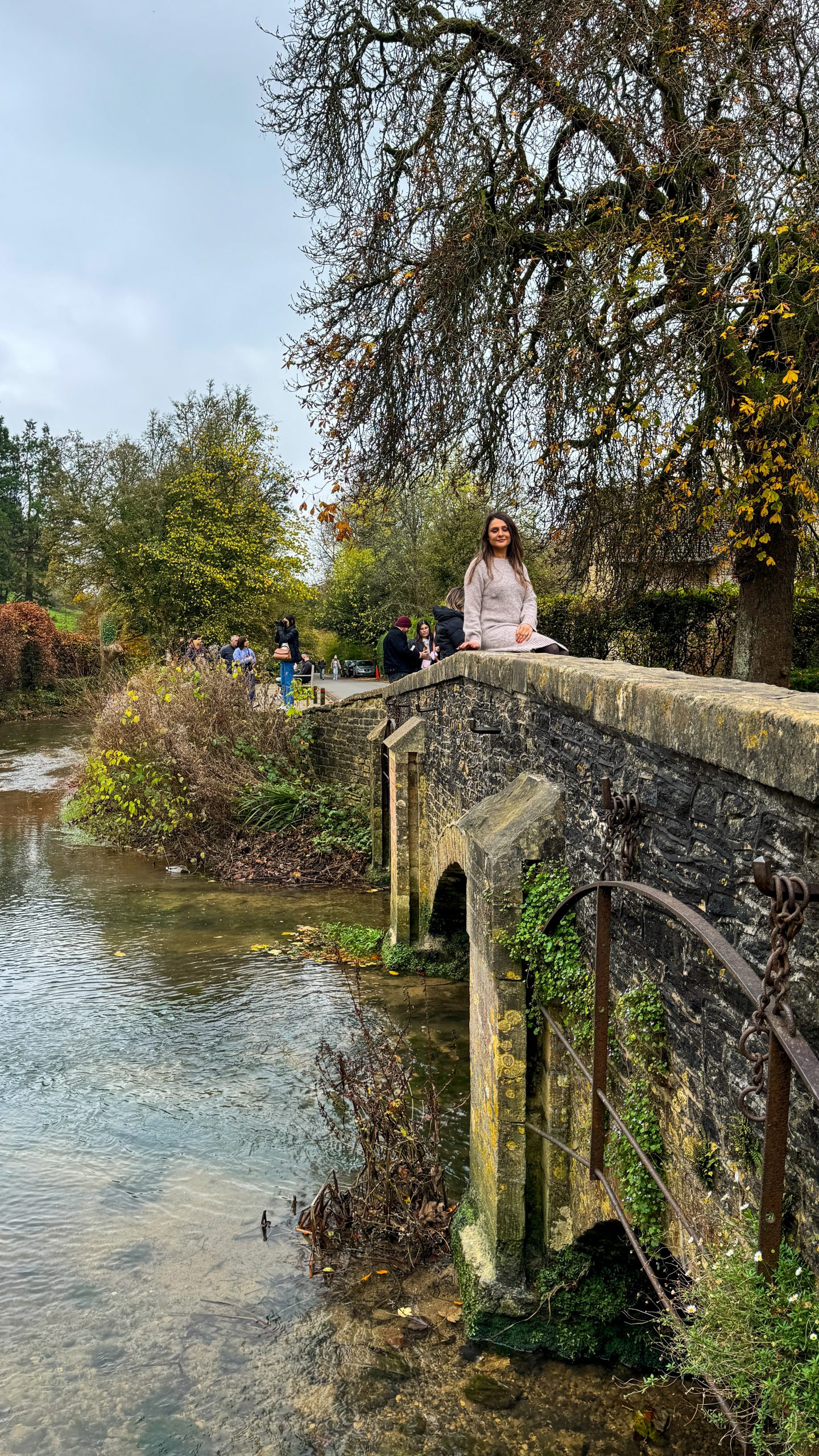 A woman sitting on the stone bridge in Castle Combe looking at the river
