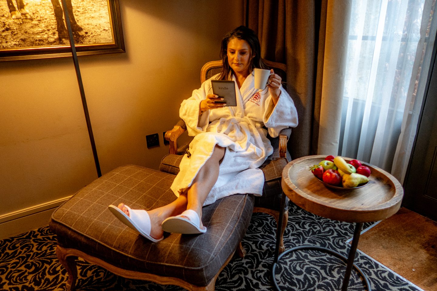 Woman sitting in a robe on a chait with a cup of tea in one hand and a kindle in the other hand at The Lygon Arms